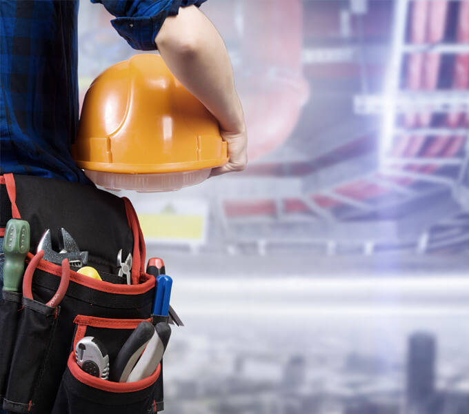 The body of a plumber/technician is cut off to the left of the picture. He is holding an orange helmet in his arms and carrying various tools around his waist. He is looking at a building site.